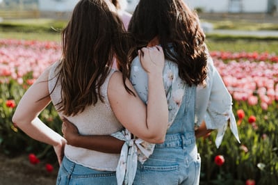 Two women from the back side-hugging in front of a field of pink and red roses