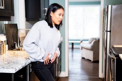 woman standing in kitchen looking troubled with hands on pelvic area and legs slightly crossed. Experiencing incontinence issues after pregnancy