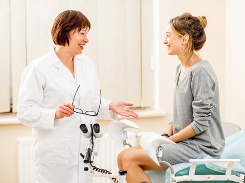A woman sitting on an examination table in a medical office, having a discussion with her OBGYN before a pelvic exam. Both individuals are engaged in a respectful and professional conversation, ensuring the patient's comfort and informed consent.